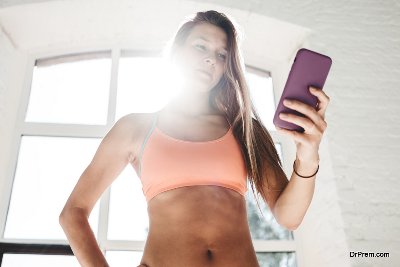 Tired sporty woman with headphones looks at smartphone and counts up spent calories after training. Fit female athlet warming up before workout and hold smartphone in hand early morning in gym